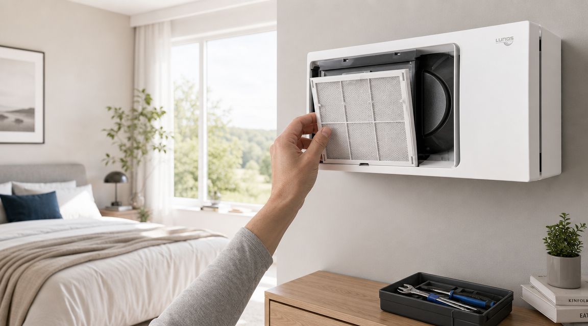 En person udskifter filteret i et Lunos ventilationsanlæg monteret på væggen i et moderne lyst soveværelse.