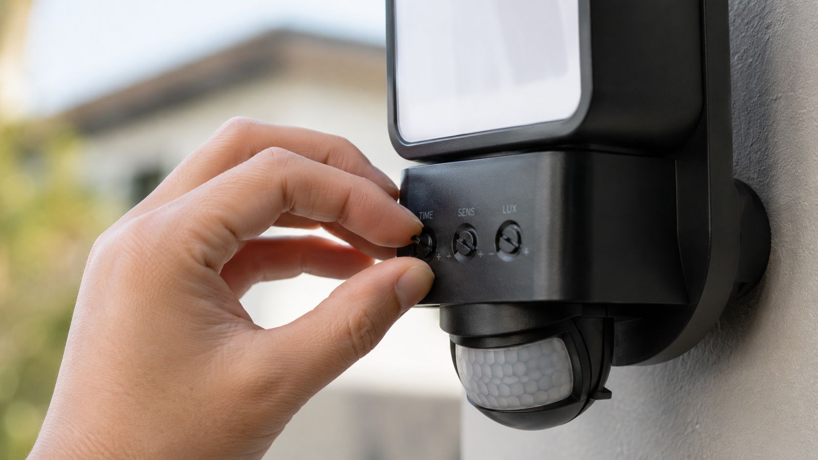 En hånd justerer indstillingerne på en udendørs lampe med bevægelsessensor monteret på en husmur.